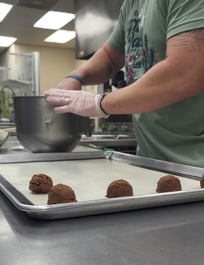 Person in a green T-shirt wearing gloves forming chocolate cookie dough balls on a baking sheet in a commercial kitchen.