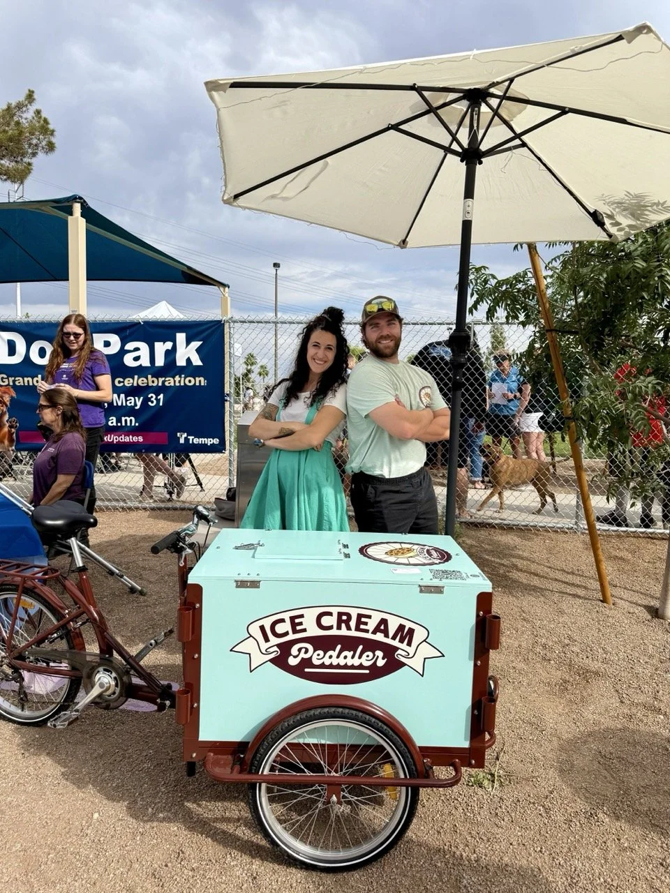 A man and a woman standing behind a light blue ice cream bike cart with a sign that says "Ice Cream Pedaler". They are under a large white umbrella at an outdoor event, with other people and dogs in the background.