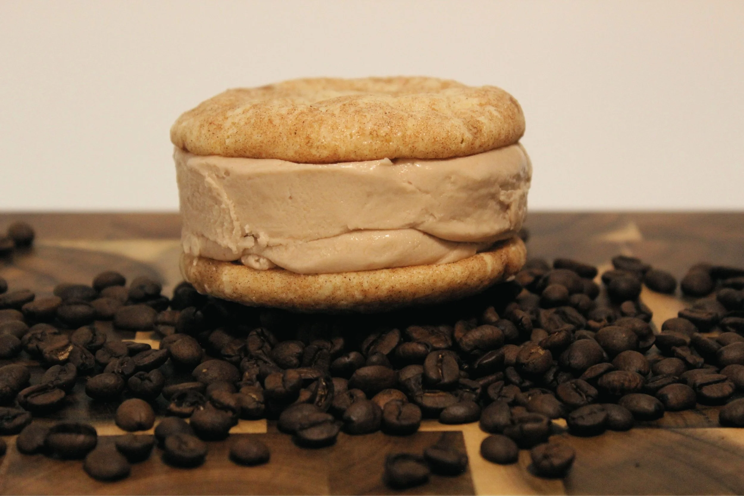 A snickerdoodle cookie ice cream sandwich with coffee ice cream, surrounded by scattered coffee beans, on a wooden surface with a plain white background.