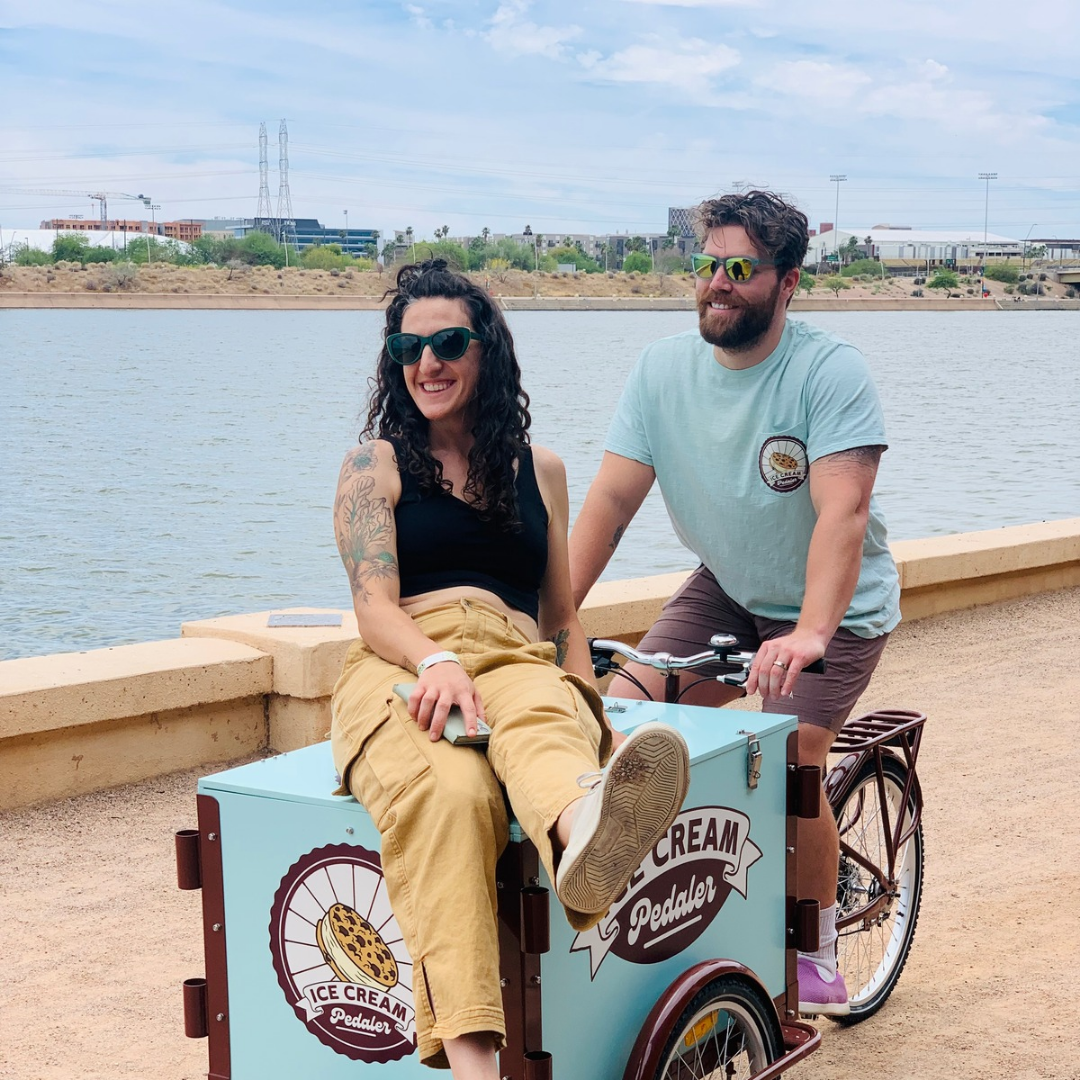 A woman and man smiling with sunglasses by a river. The woman is sitting in an ice cream bicycle cart, and the man is riding the bicycle. The cart has a logo that says "Ice Cream Pedaler."