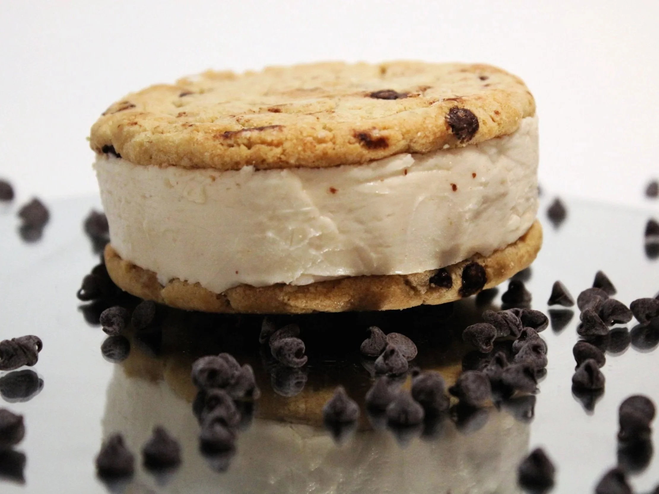 A close-up of a dessert with a chocolate chip cookie top layer, a middle layer of vanilla ice cream, and a cookie base, surrounded by scattered mini chocolate chips on a reflective surface.