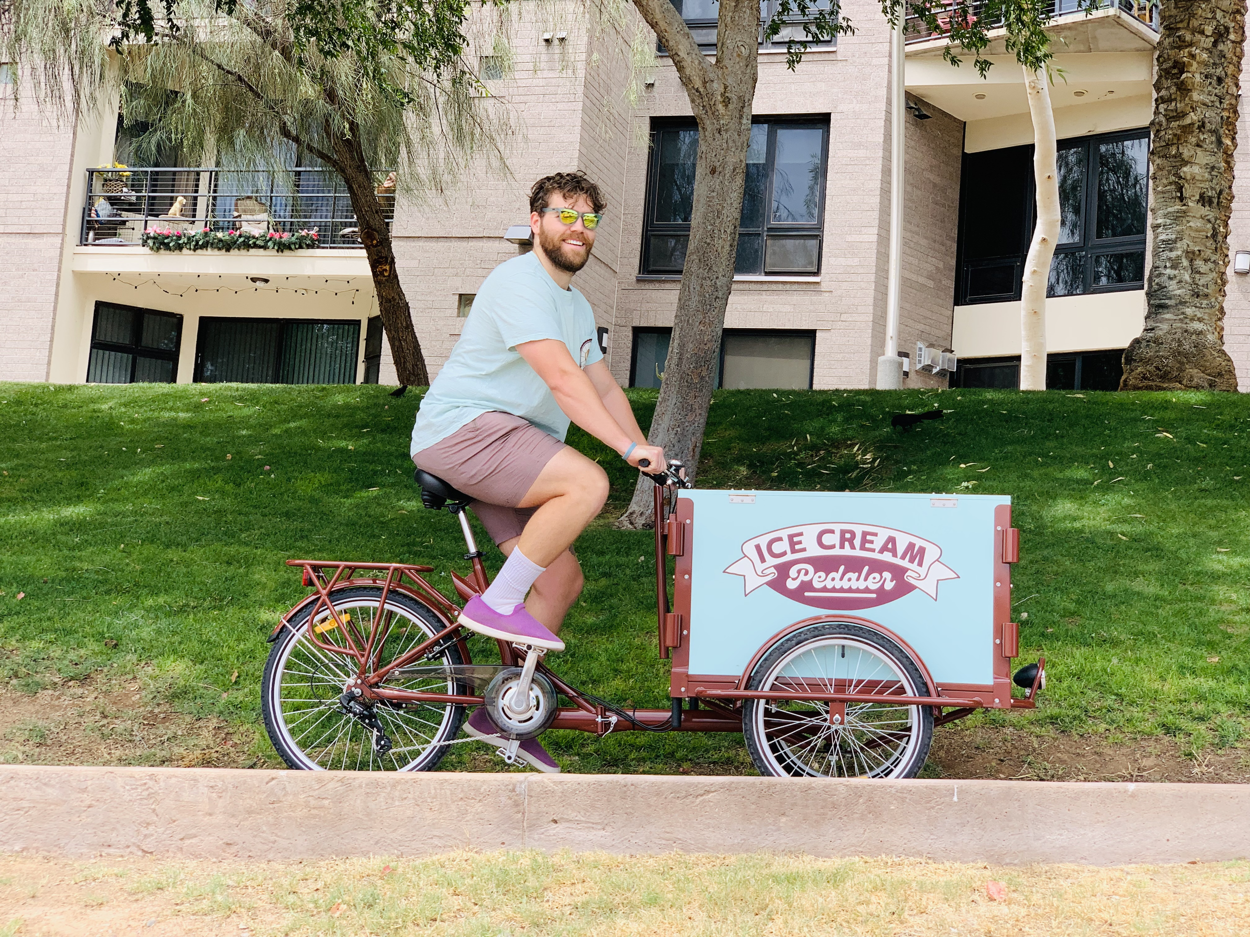 A man with a beard and sunglasses riding a red ice cream pedal bike in a grassy area next to a building with large windows and trees.
