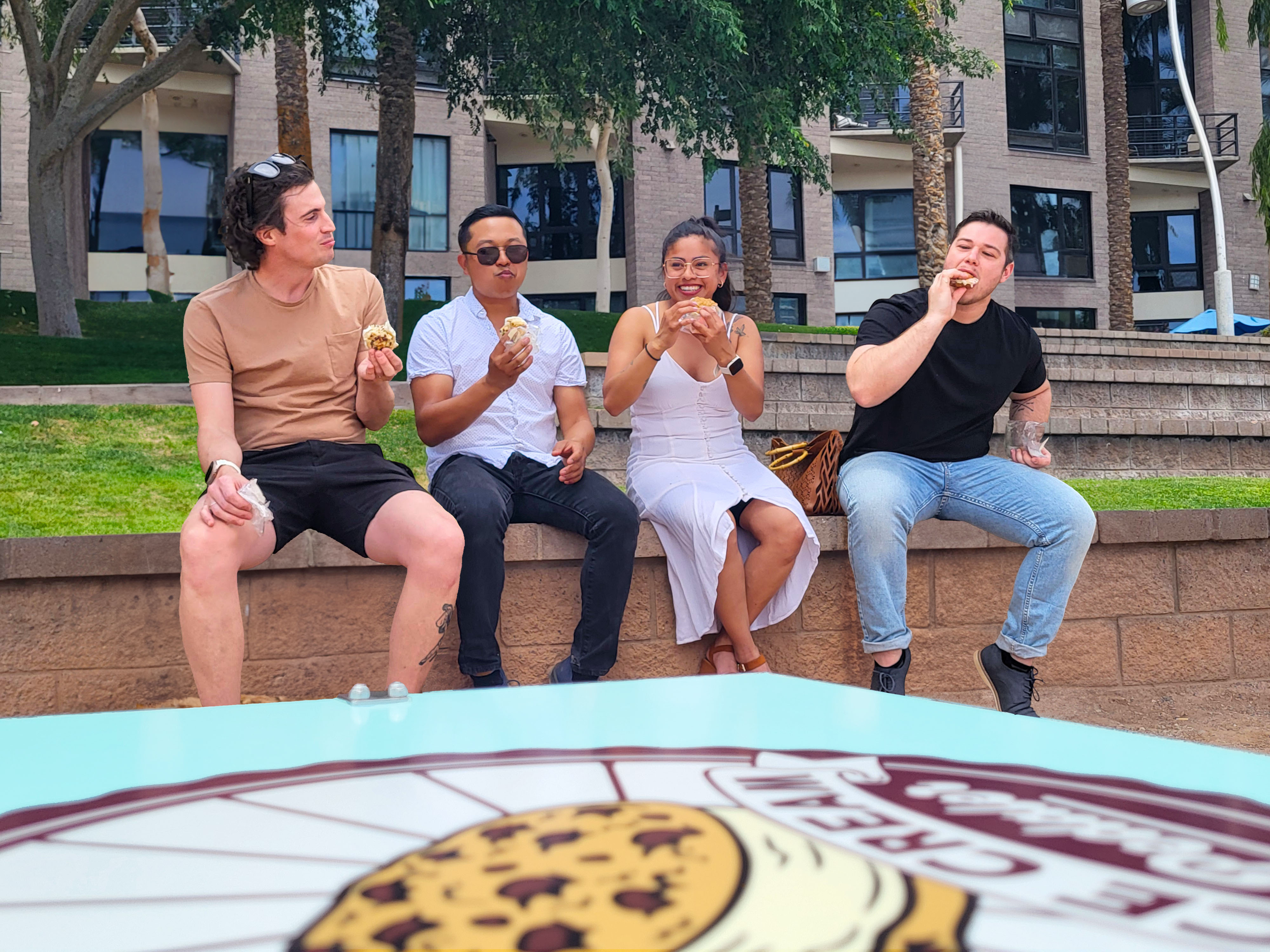 Four friends sitting outdoors on a brick wall, eating cookies and smiling, with trees and a modern building in the background.