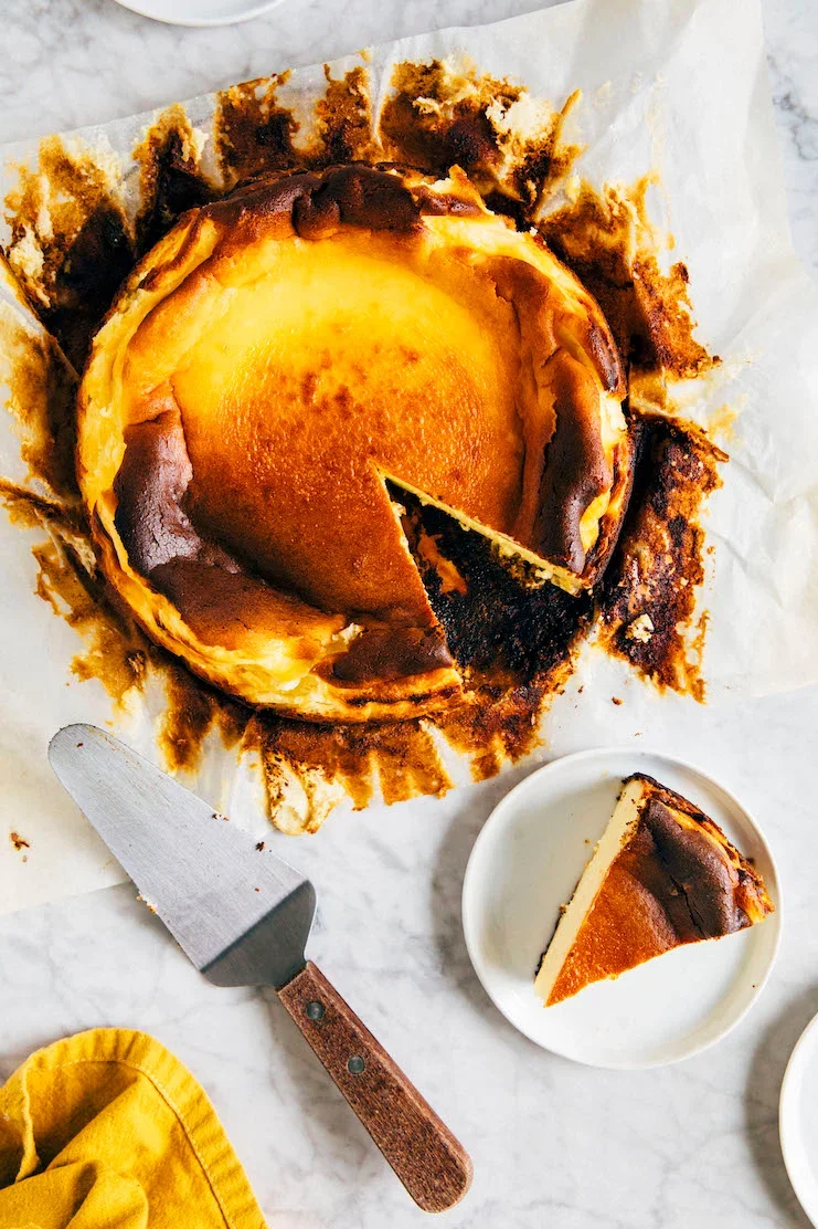 Baked cheesecake with a burnt top, with a slice cut out and placed on a white plate, a cake server, and a yellow cloth nearby.