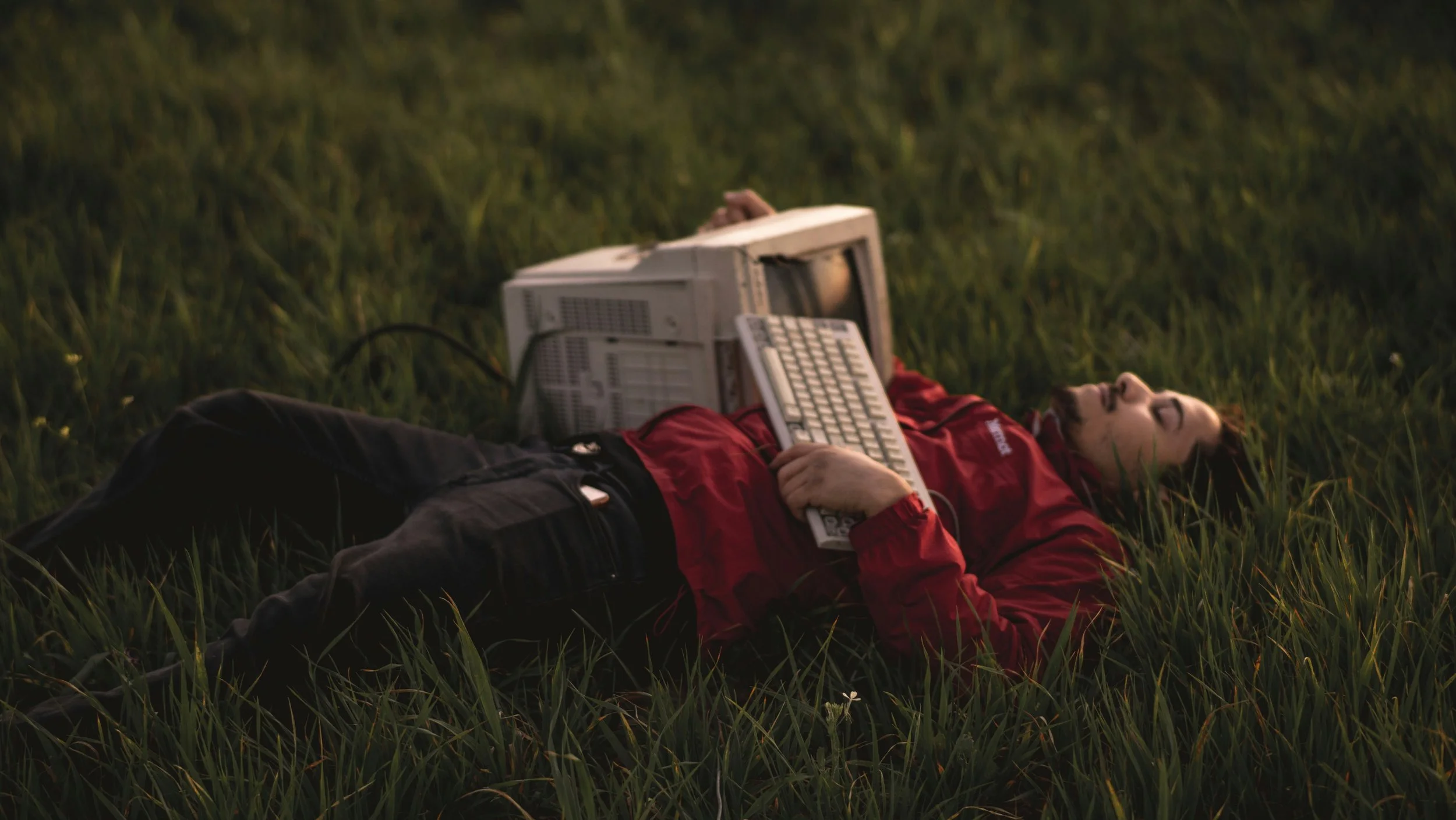 A person lying on their back in a grassy field with closed eyes, holding a keyboard on their chest and a computer tower beside them.