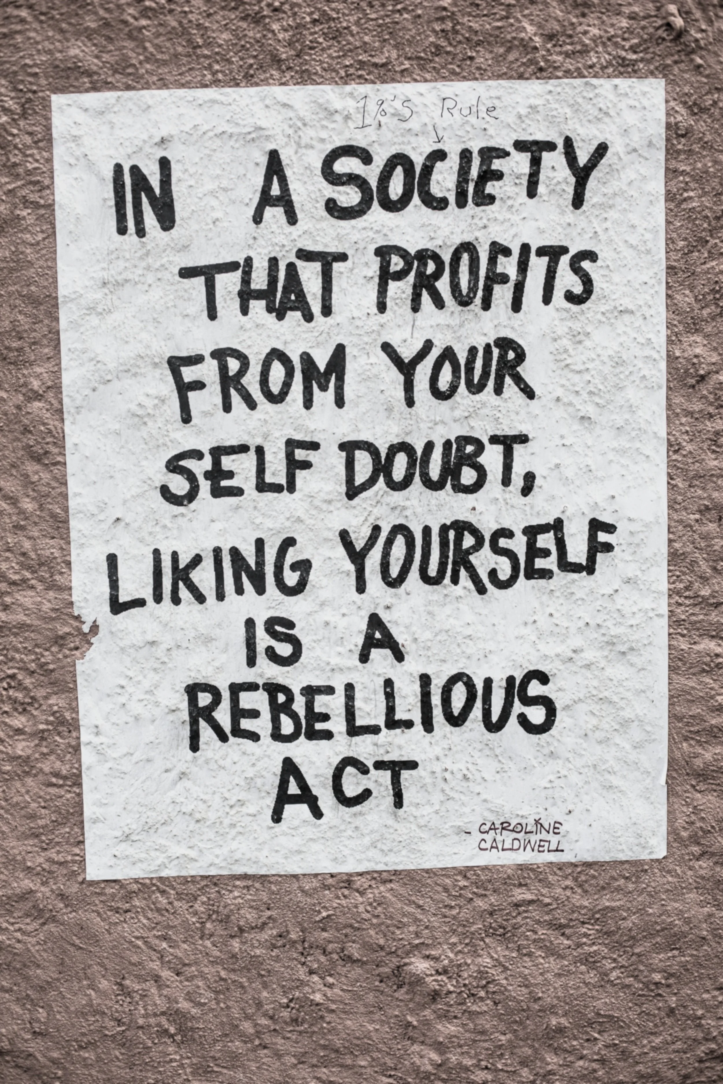 A handwritten quote on paper, titled '1955 Rule,' stating 'In a society that profits from your self doubt, liking yourself is a rebellious act' by Caroline Caldwell, displayed on a textured wall.