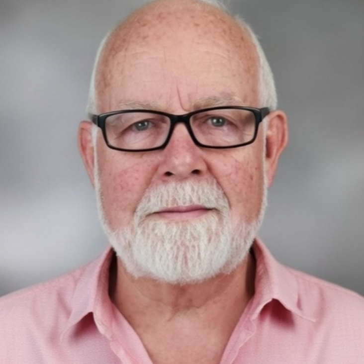 Close-up portrait of an older man with a white beard and glasses, wearing a pink collared shirt, against a plain gray background.