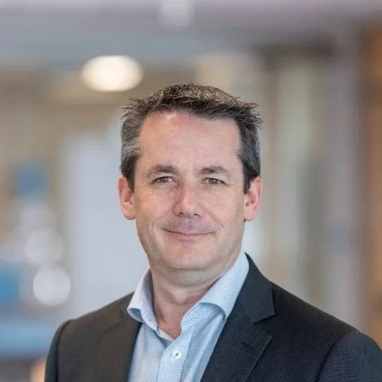 A professional man with dark hair and a light blue shirt, smiling in a business setting.