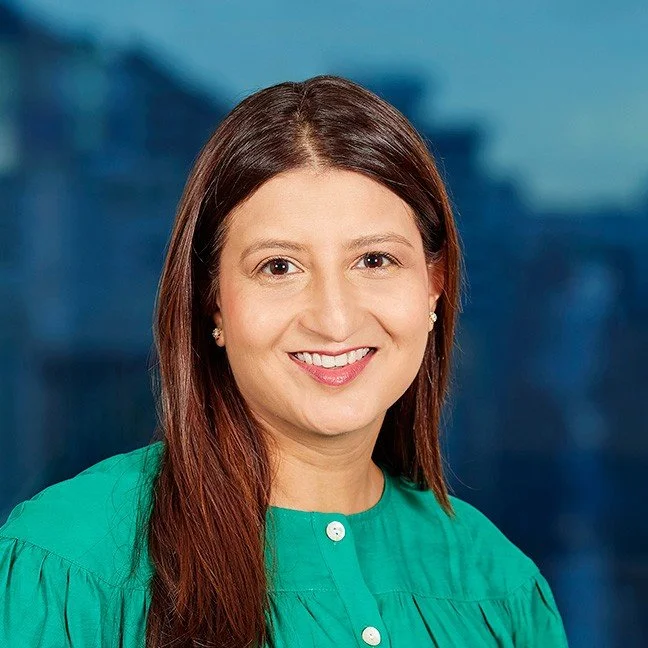 A woman with long brown hair smiling, wearing a green blouse and small earrings, against a cityscape background.