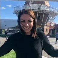 A woman with dark hair smiling, standing outdoors in front of a building with a modern glass structure, on a sunny day.