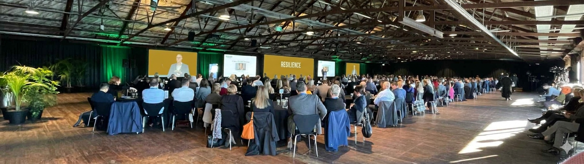 Large conference hall filled with attendees sitting at round tables listening to a speaker on stage. The stage displays screens with the word 'RESILIENCE.' The hall has a high, exposed ceiling with lighting, greenery on the left side, and some people seated or standing on the right.