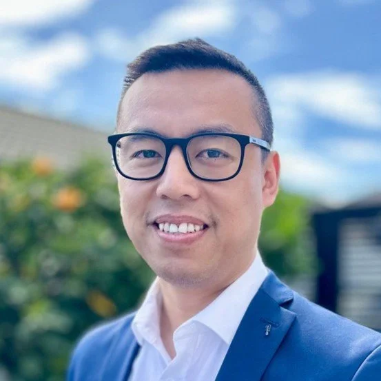 Portrait of a man wearing glasses and a navy blue suit jacket, standing outdoors with a blurred background of green foliage and a cloudy blue sky.