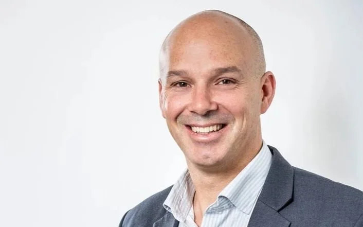 A smiling man with a shaved head, wearing a dark suit jacket and light blue dress shirt, against a plain gray background.