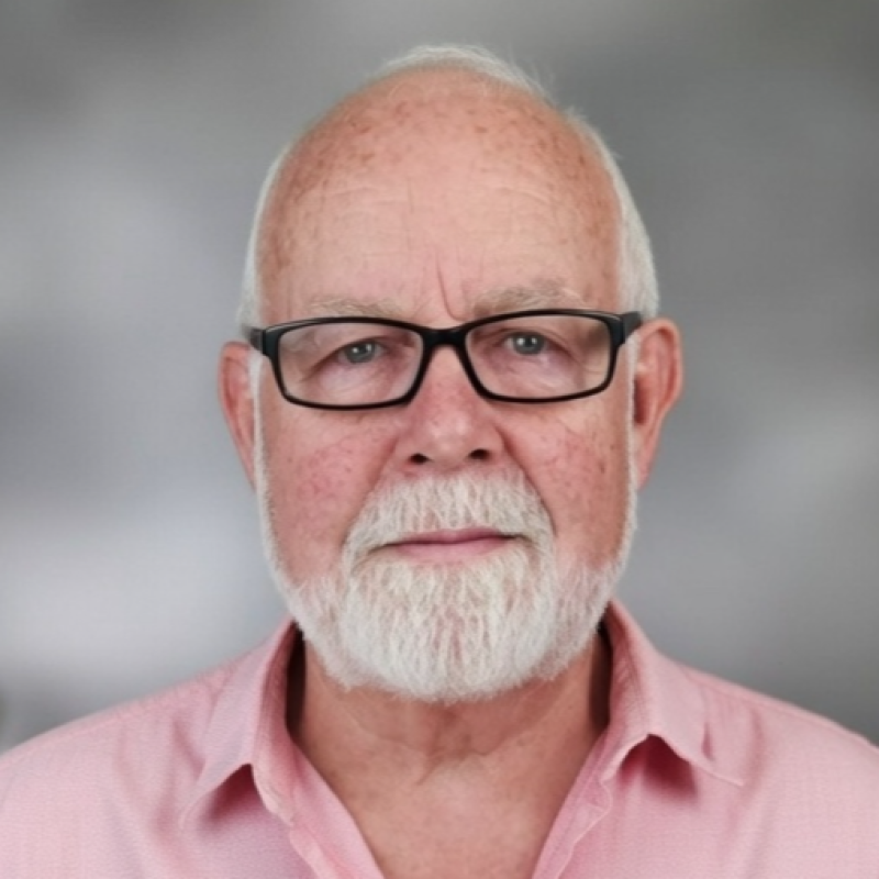 Close-up portrait of an elderly man with glasses, white beard, and mustache, wearing a pink collared shirt, against a blurred gray background.