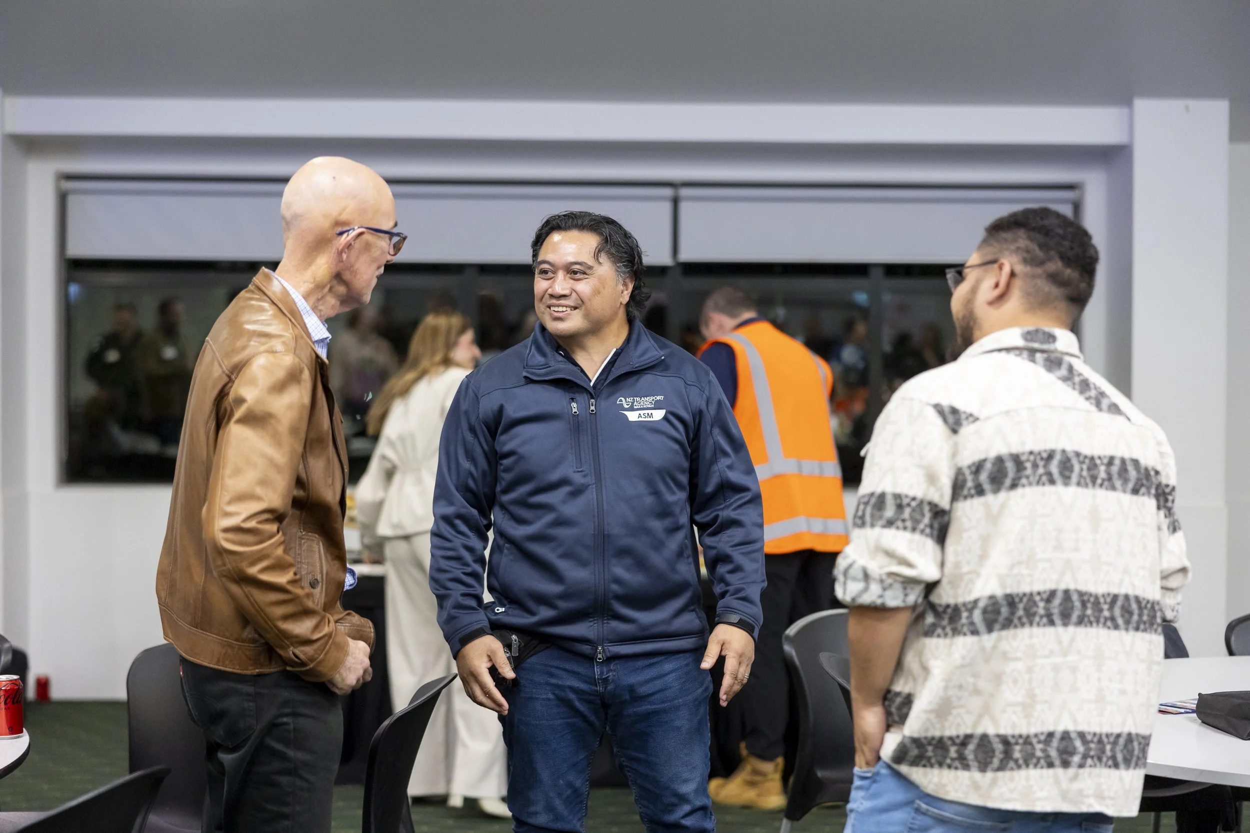 Three men engaged in conversation at an indoor event, with a woman and others in the background. One man is wearing a brown jacket, another a navy jacket with a name tag, and the third a white and black patterned shirt.