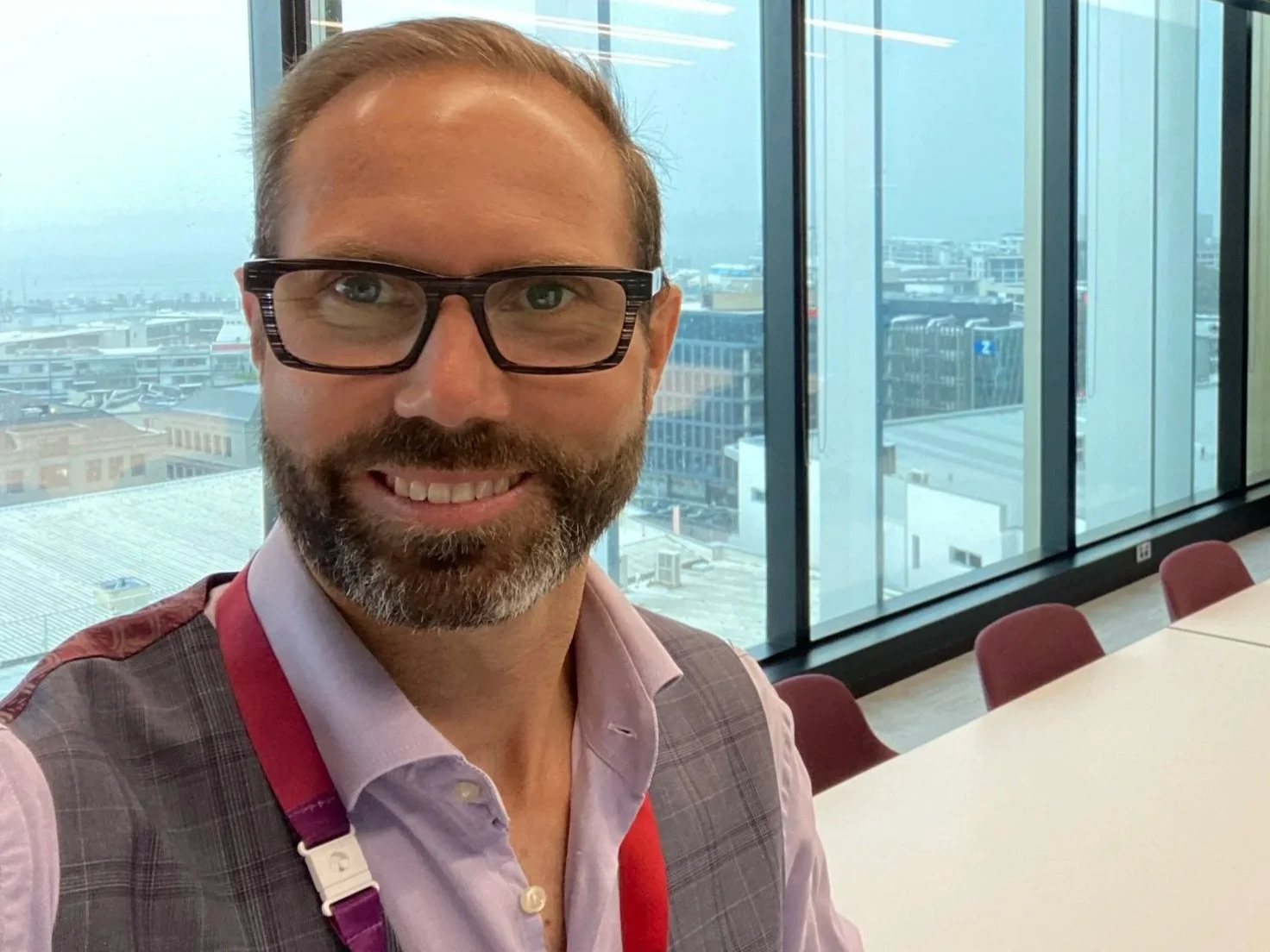 A man with glasses and a beard smiling in an office with large windows showing a cityscape behind him.