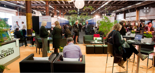 People networking and chatting at a coffee trade show or conference in a large, well-lit industrial-style space with plants and booths.