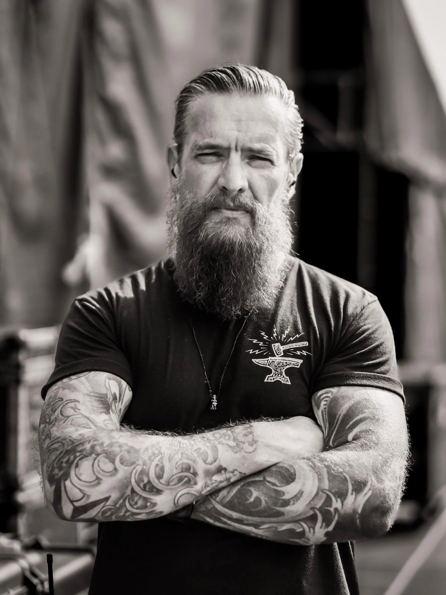 A man with a long beard and tattooed arms crossing his arms, standing on a stage with equipment in the background, wearing a black T-shirt with a skull illustration.