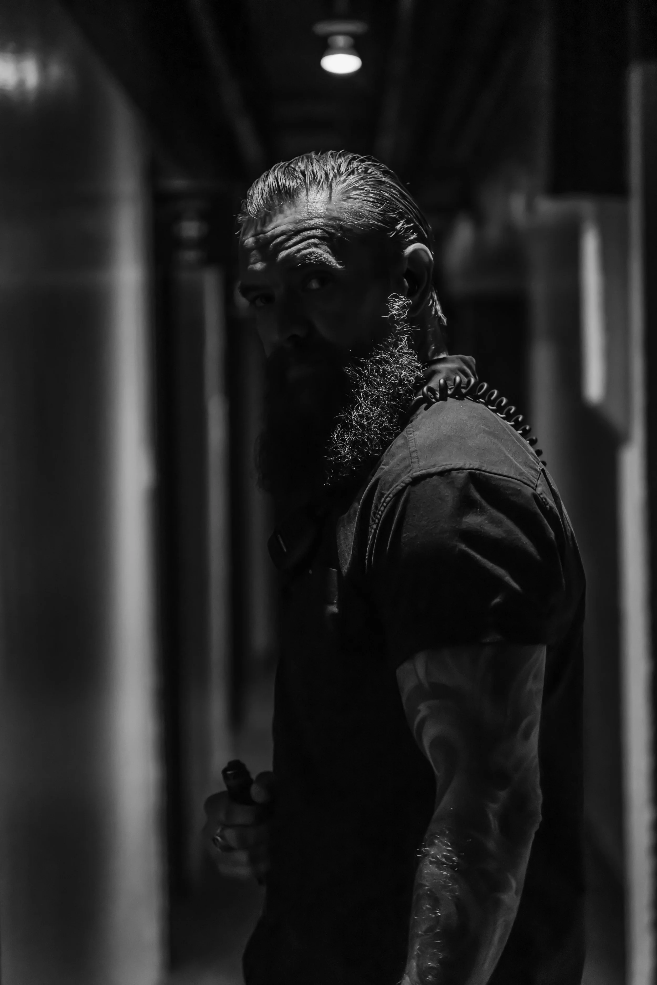 A black-and-white photo of a man with slicked-back hair and a beard, providing security for an international band, looking over his shoulder at the camera in a dimly lit corridor backstage of a major venue