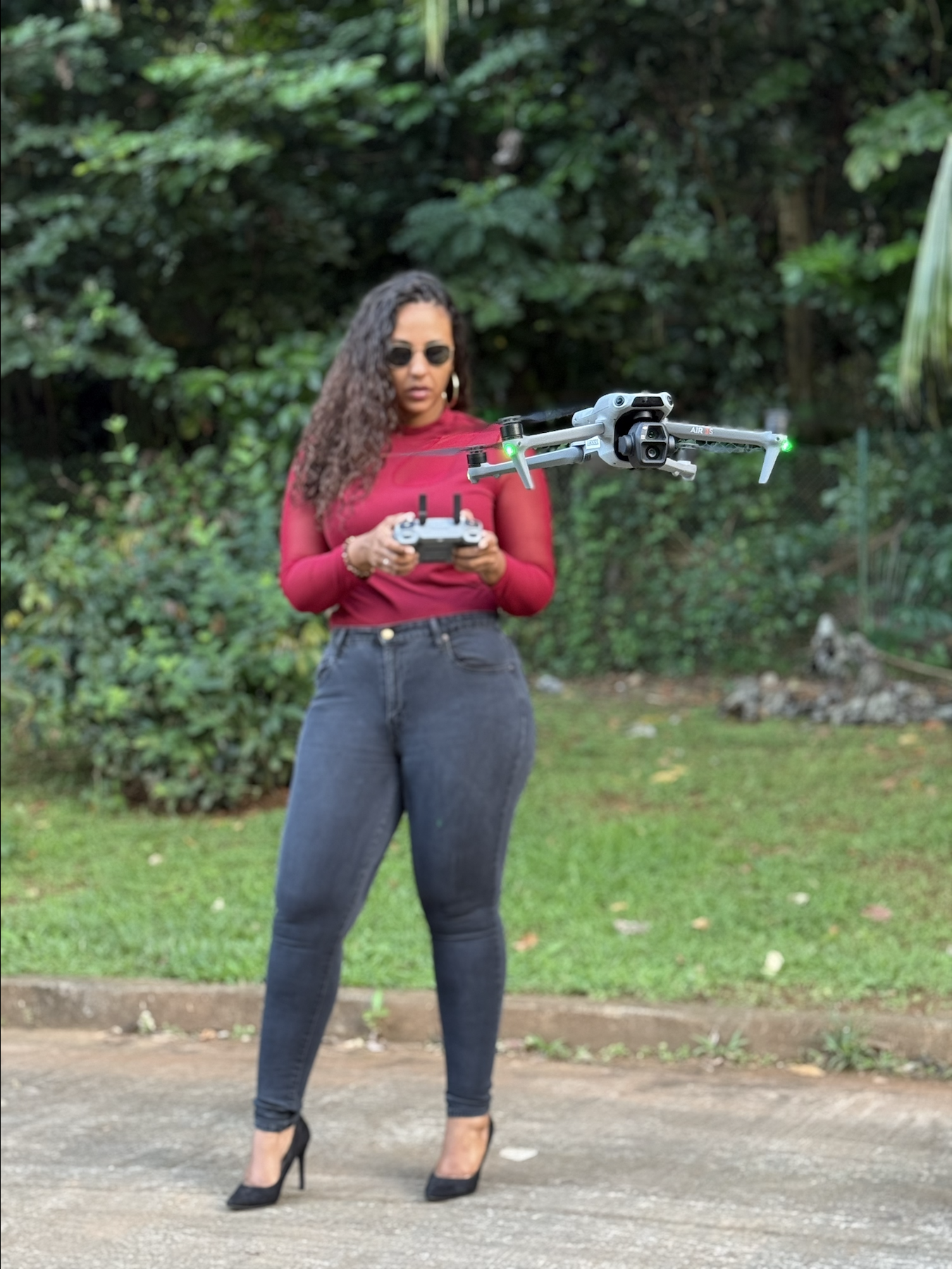 Une femme debout en extérieur, portant un haut rouge, un jean noir, des talons noirs, et des lunettes de soleil, contrôlant un drone dans un environnement verdoyant.