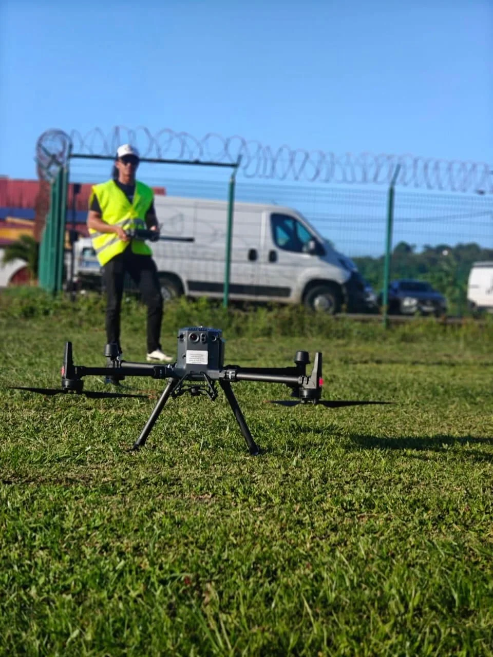 Un drone posé sur l'herbe devant une personne en gilet jaune, qui tient un contrôleur de drone, en arrière-plan un véhicule blanc près d'une clôture avec du fil de fer barbelé et un ciel bleu.