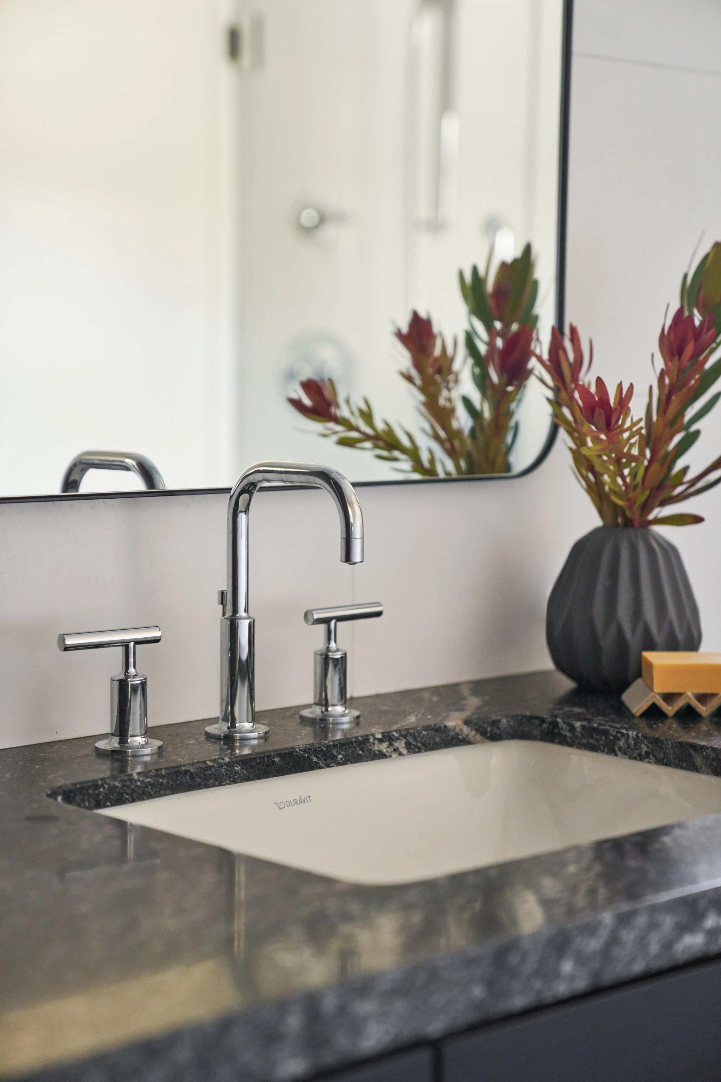 Close-up of a modern bathroom sink with a sleek faucet, a mirror, and a vase with reddish-green foliage on a gray countertop.