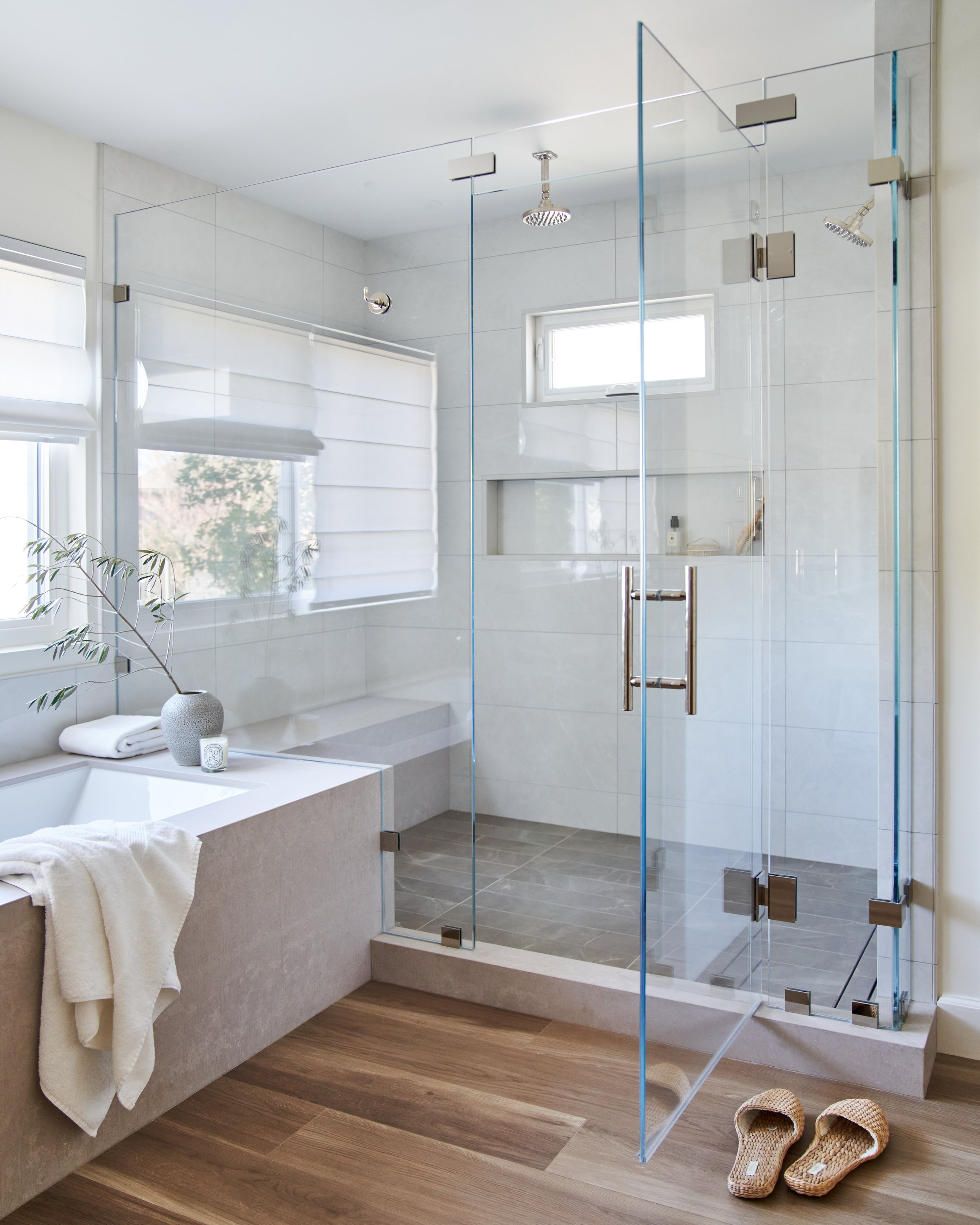 Modern bathroom with a walk-in shower enclosed by glass, a built-in bathtub with towels, and wooden flooring. There are small slippers on the floor near the shower.