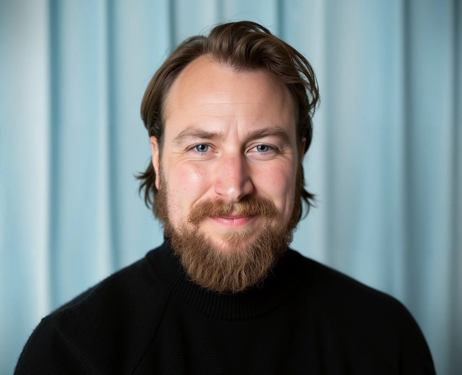 Portrait of a smiling man with blue eyes, light skin, brown hair, and a beard, wearing a black sweater, against a blurred blue background.