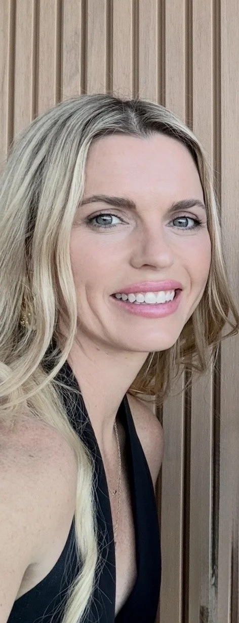 A woman with blonde wavy hair and blue eyes smiling, wearing a black sleeveless top, standing in front of a wooden paneled background.