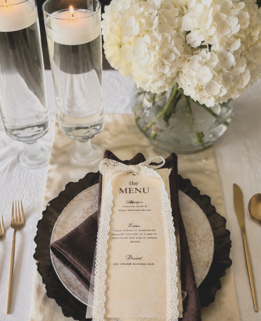 Elegant table setting featuring a menu card with lace trim, gold flatware, two tall glasses of water, a lit candle, and a large glass bowl of white hydrangea flowers.