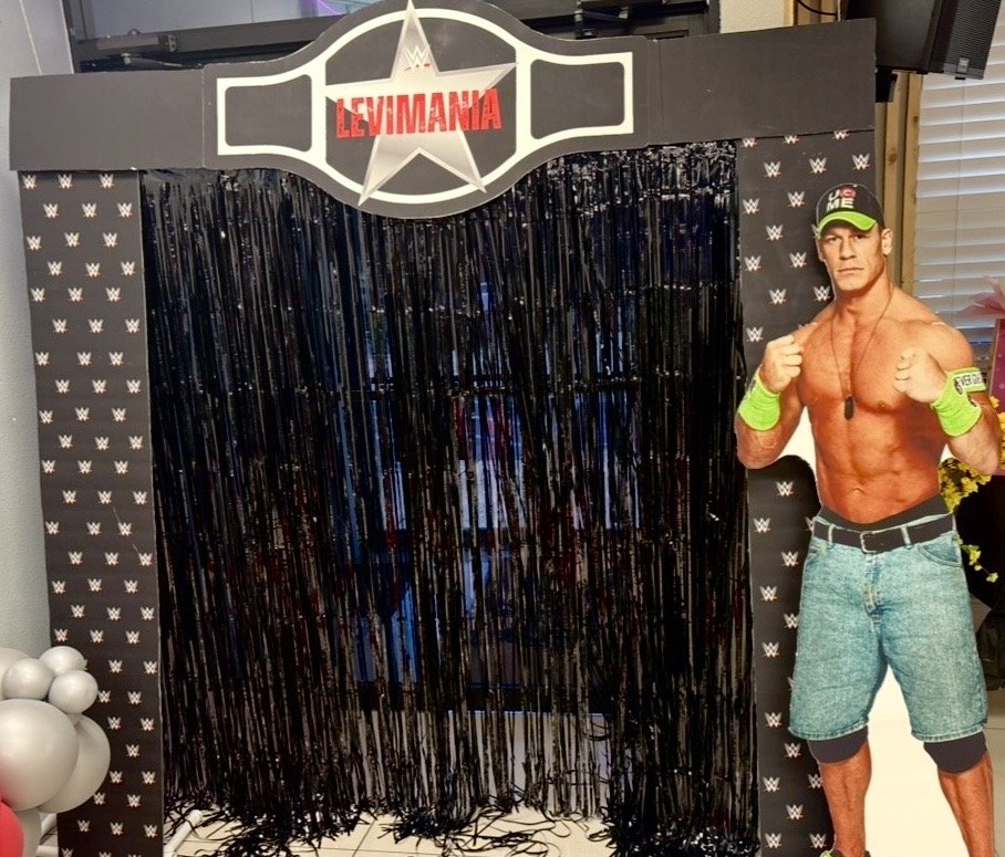 personalized wrestling event display with a curtain backdrop, WWF logo patterned sides, a sign at the top, and a life-sized cardboard cutout of a shirtless wrestler posing with clenched fists, wearing a cap, wristbands, jeans, and a weight belt.