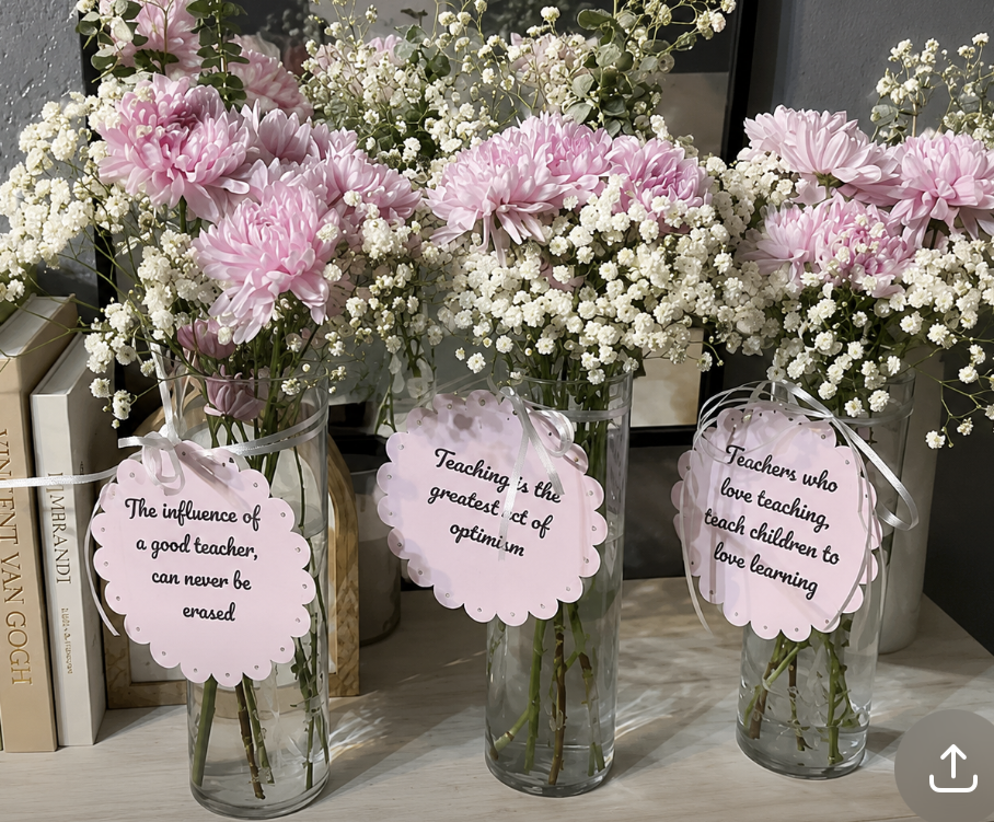 Three vases with pink chrysanthemums and white baby's breathe flowers, each with a pink tag featuring a quote about teaching, placed on a light-colored surface with books behind them.