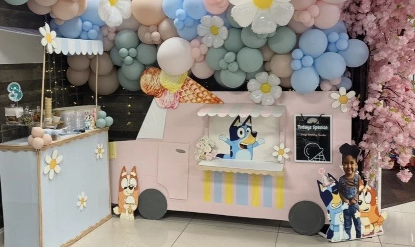 A colorful, bluey-themed ice cream truck display decorated with pastel-colored balloons, flowers, and cartoon characters from the 'Bluey' TV show, with a cardboard cutout posing next to bluey and bingo.