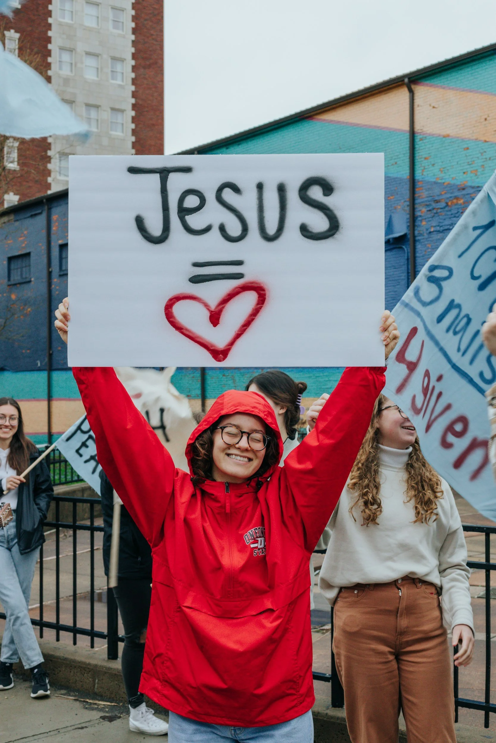 A woman in a red jacket and glasses is holding a sign that says 'JESUS = love' with a heart symbol on it. She is smiling at a group of people participating in a street protest or rally, with others holding signs around her. The background shows urban buildings and a colorful mural.