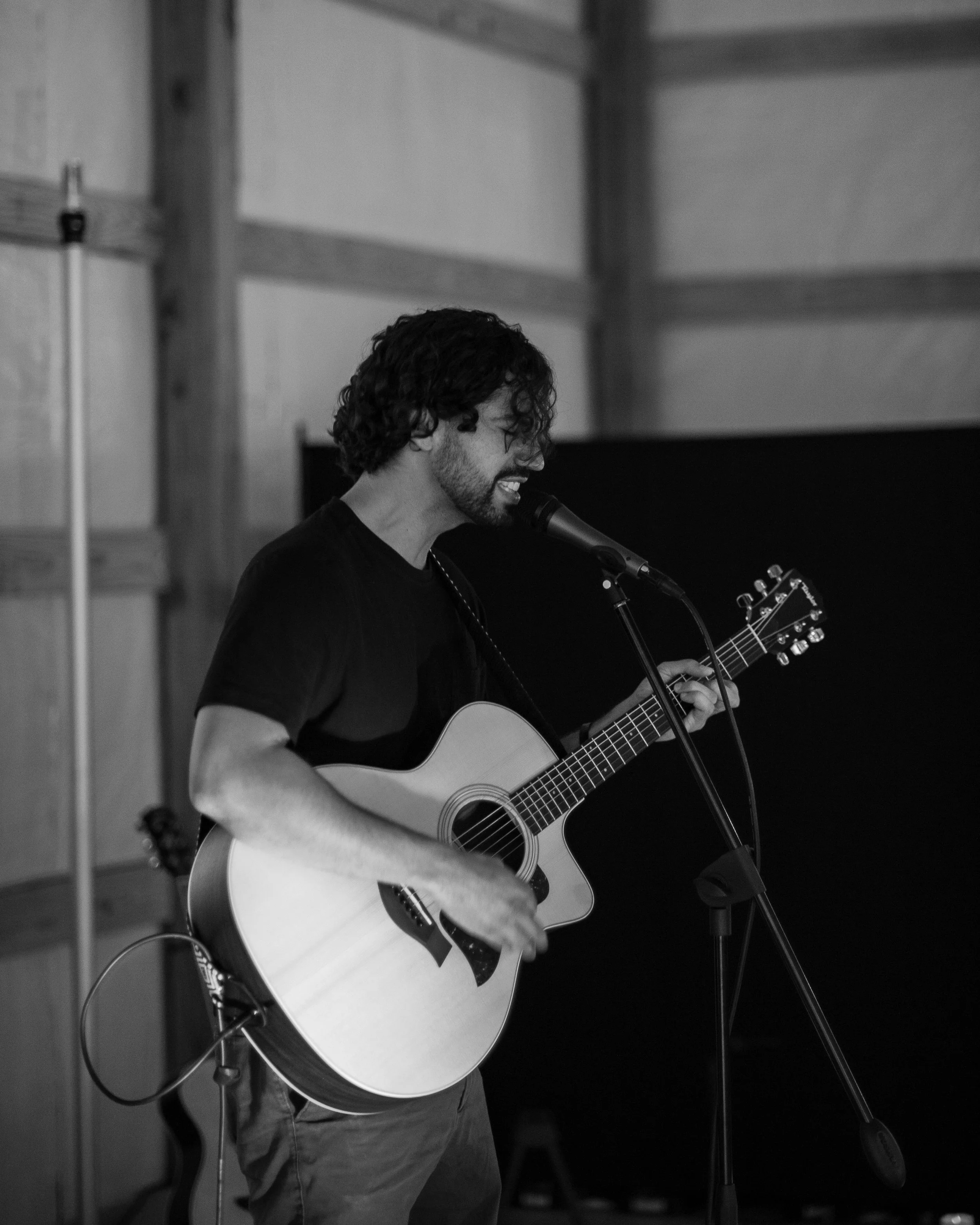 A man with curly hair and glasses playing an acoustic guitar and singing into a microphone in a room with wooden wall paneling.
