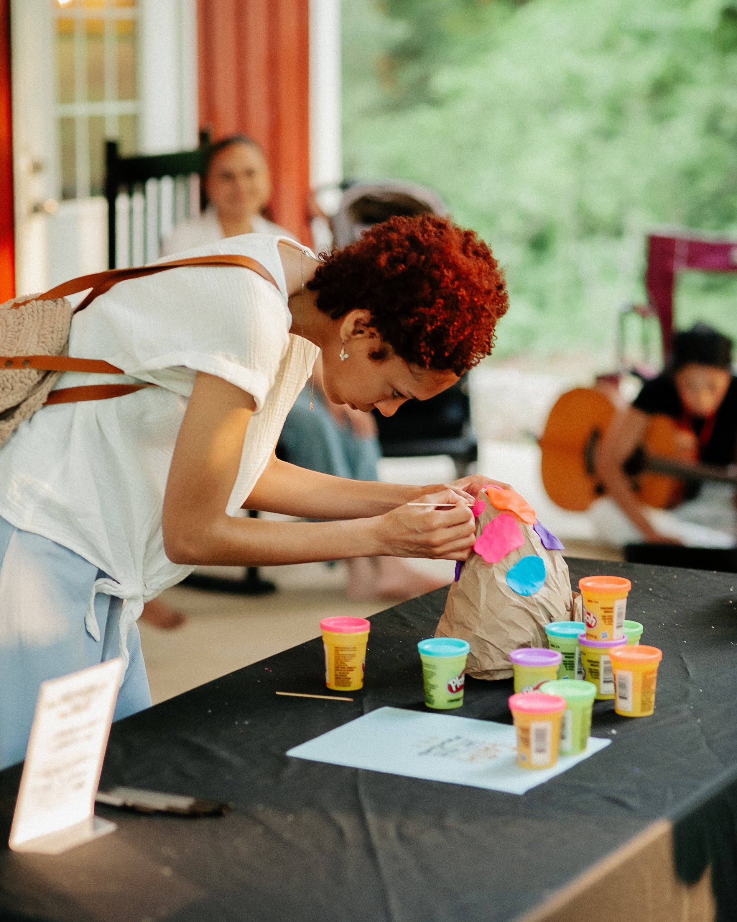 A woman with curly red hair in a white shirt is bent over, working on a craft project with colorful Play-Doh on a black table. In the background, there are other people, a woman with dark hair, a person in an orange shirt, and a girl with glasses. The setting appears to be an outdoor or semi-outdoor space with a green, wooded backdrop and a red structure.