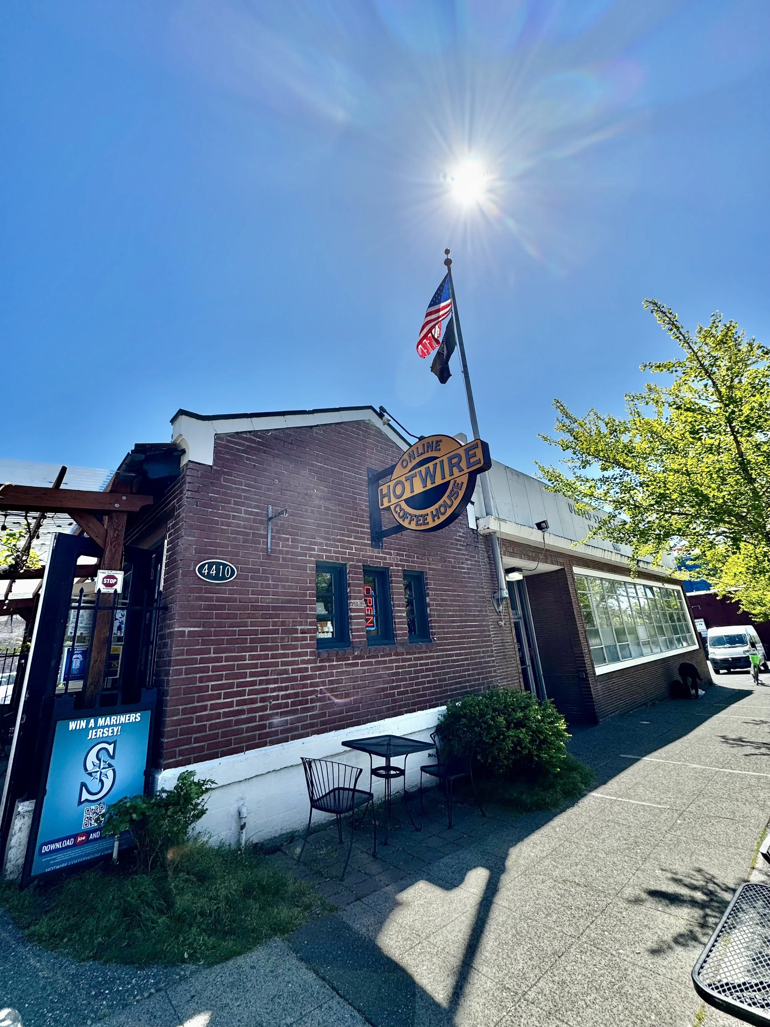 The exterior of a restaurant called Hotwire Coffee House with a red brick building, a sign, an American flag, and outdoor seating, trees, and a clear sunny sky.