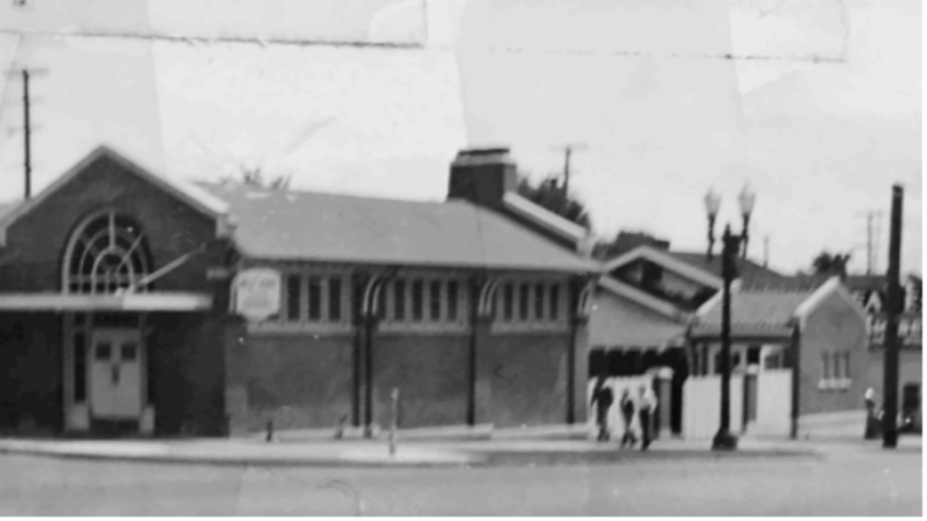 Black-and-white photo of a small brick building with a peaked roof and a large circular window, with people walking on the sidewalk.