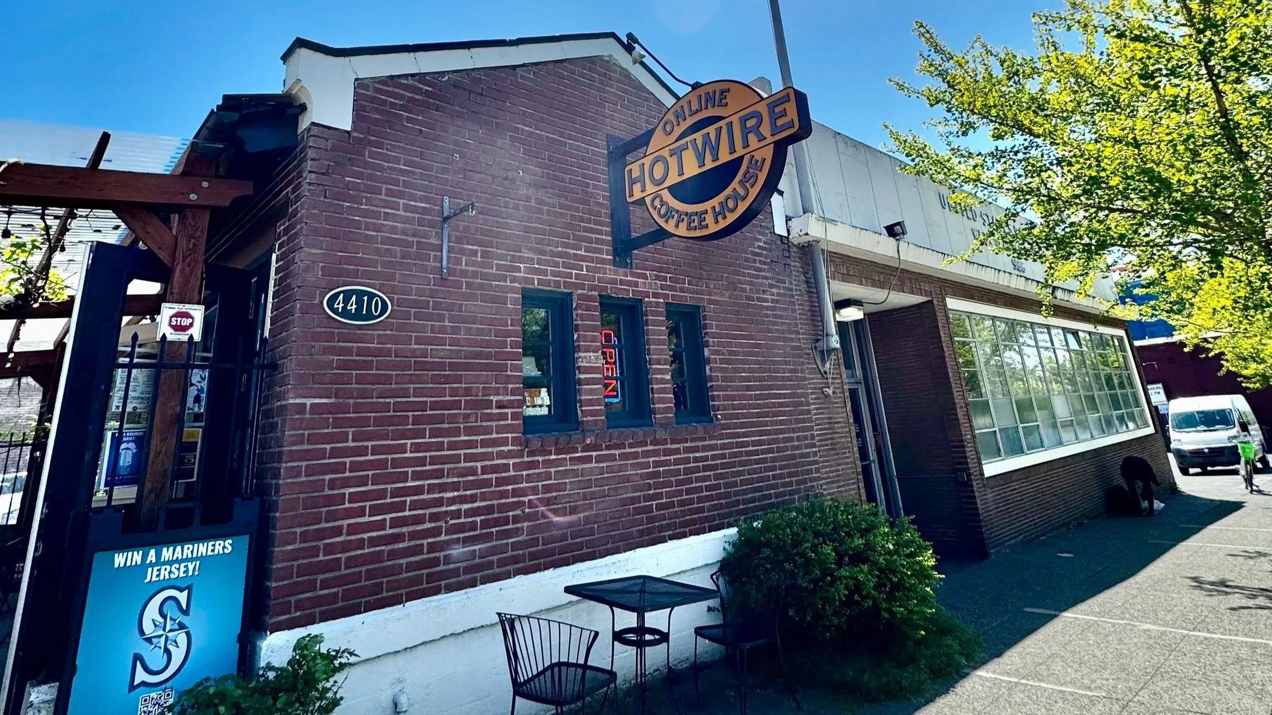 Exterior of a brick storefront with a sign that reads 'Online Hotwire Coffee House.' There is a small table and chairs outside, and a sign that says 'Win a Mariners Jersey!' with a QR code. A stop sign is visible near the entrance.