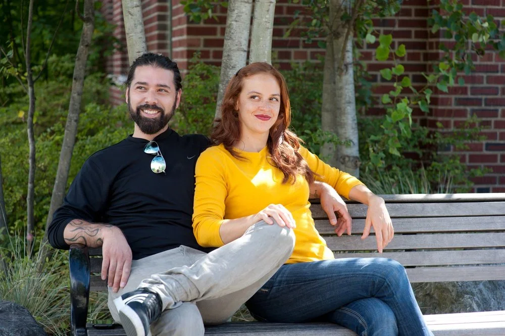 A man and woman sitting on a park bench, smiling, with trees and a brick building in the background.