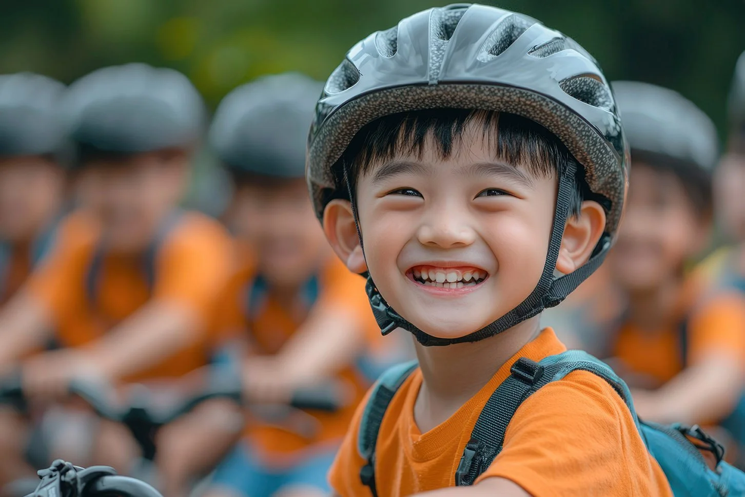 Young boy wearing a bicycle helmet and orange shirt, smiling happily at a group of children also in helmets and orange shirts, outdoors in a green setting.