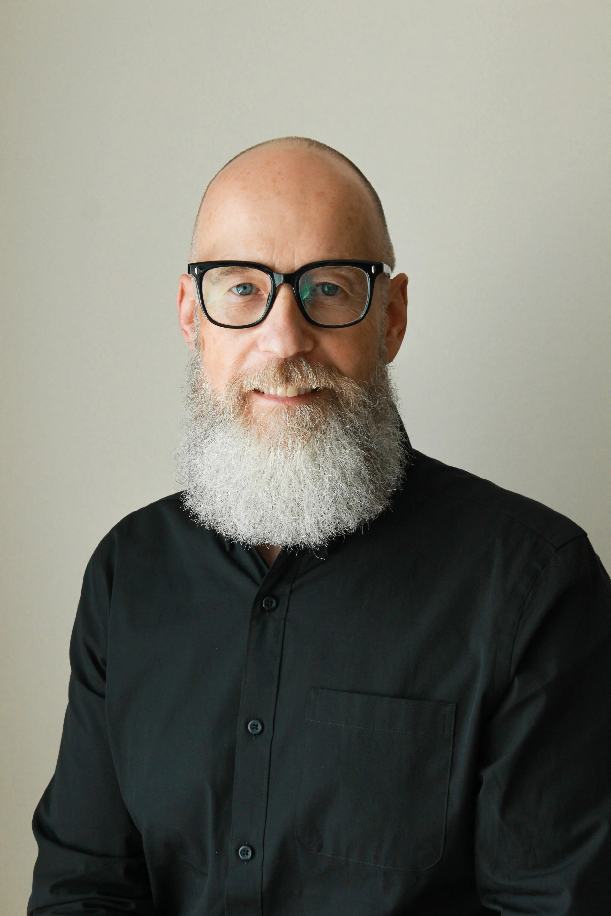 A middle-aged man with a full beard and glasses, wearing a black shirt, smiling and posing against a plain light colored wall.