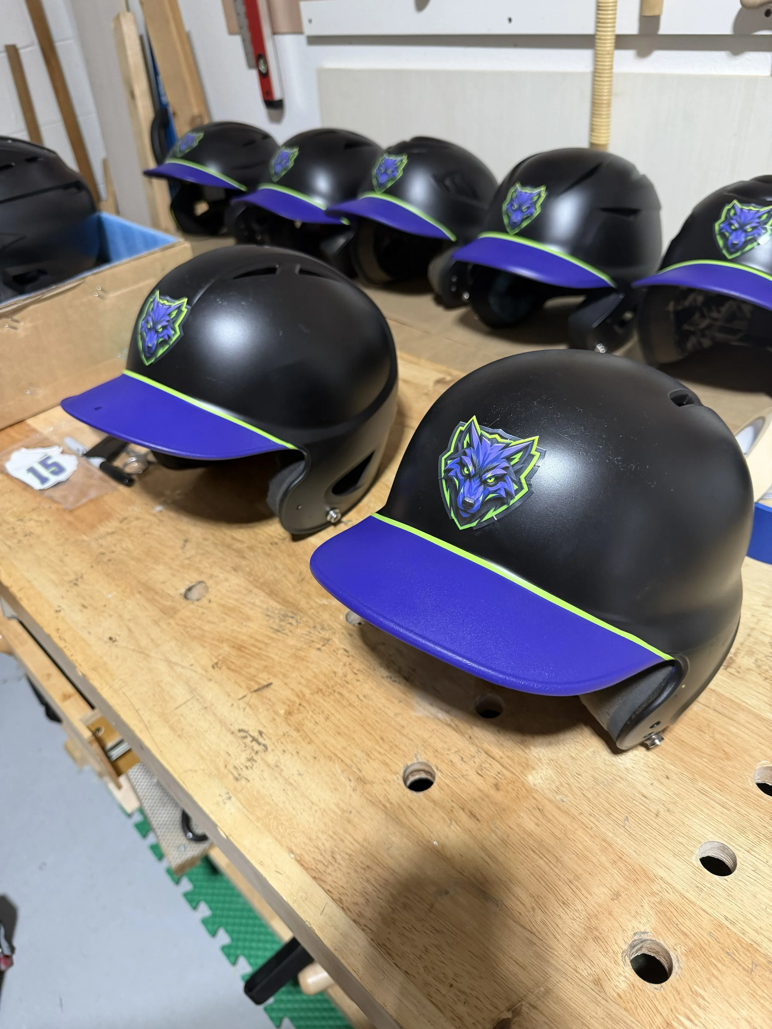 Black helmets with blue and green wolf logo, lined up on a wooden workbench.