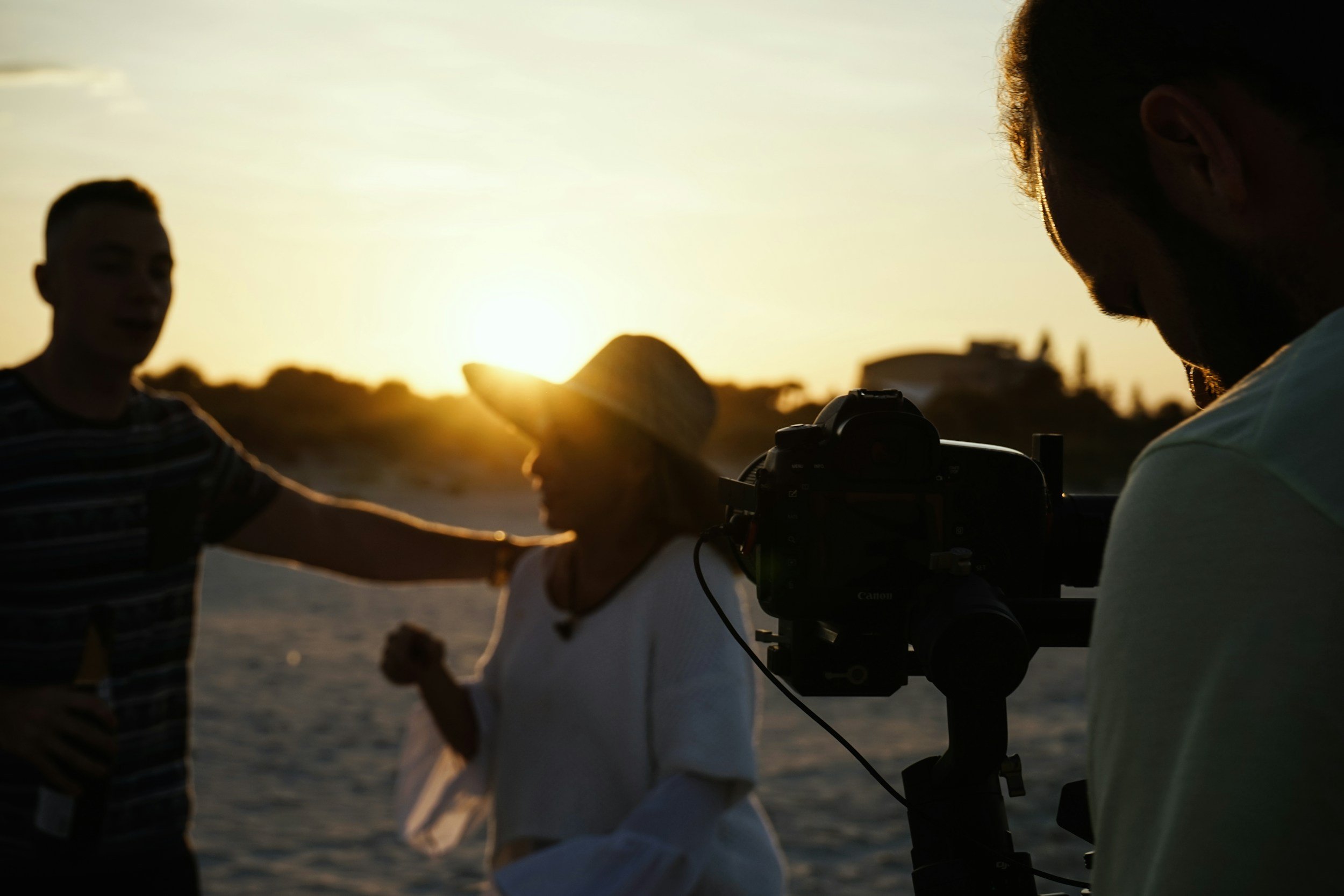 Silhouettes of three children interacting outdoors during sunset, one appears to be taking a photo with a camera.