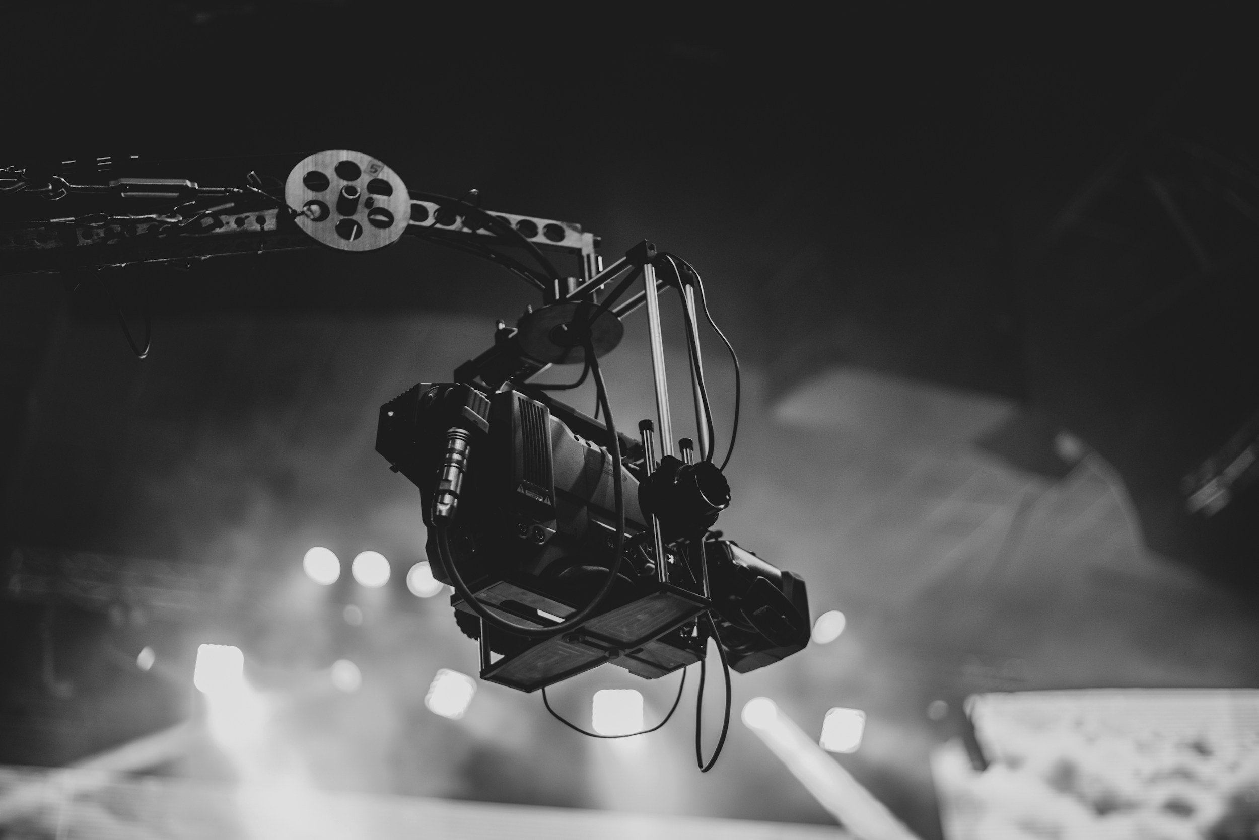 Black and white photo of a professional camera crane or jib, used for filming, suspended in the air against a blurred background of lights.