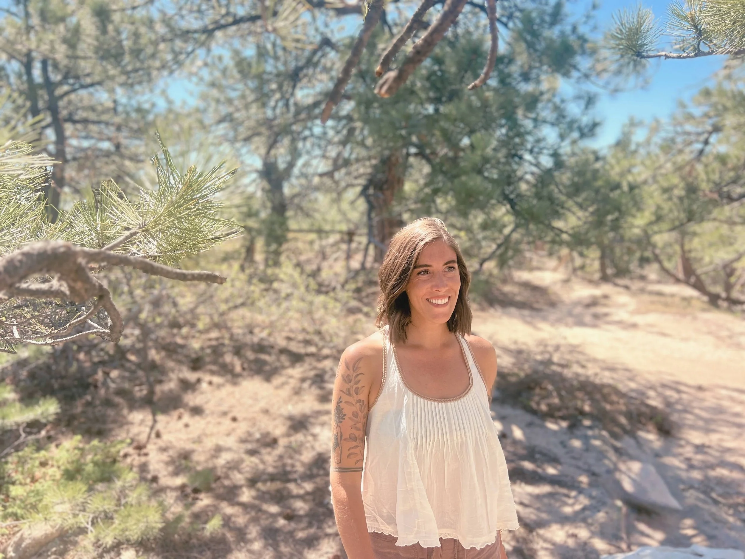 A woman with brown hair, tattoos on her arms, wearing a white sleeveless dress, smiling, standing outdoors in a sunny forested area with trees and blue sky in the background.
