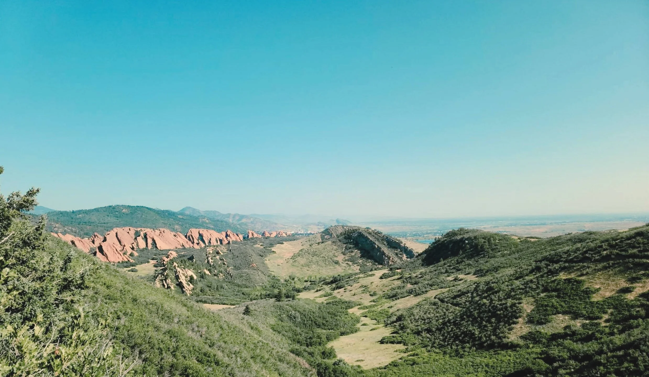 Scenic view of Boulder, Colorado with rolling green hills, rocky red formations, and a distant river under a clear blue sky.