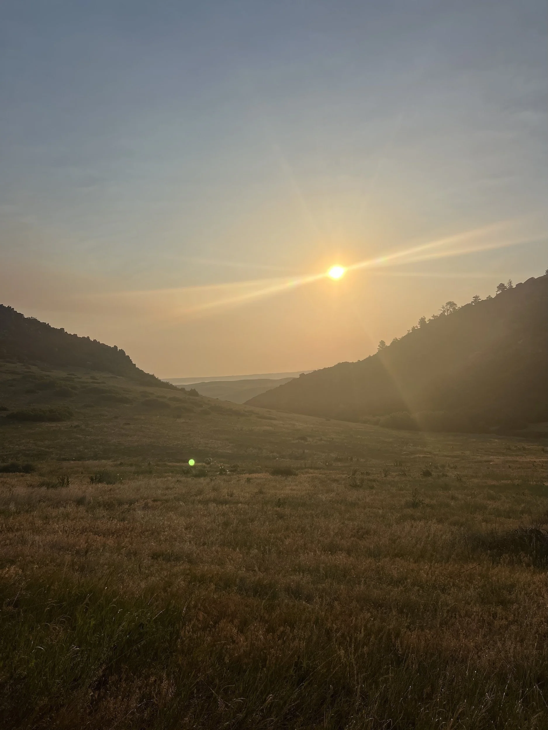 Sunset over a valley with grassy fields and rolling hills in the distance, under a partly cloudy sky.