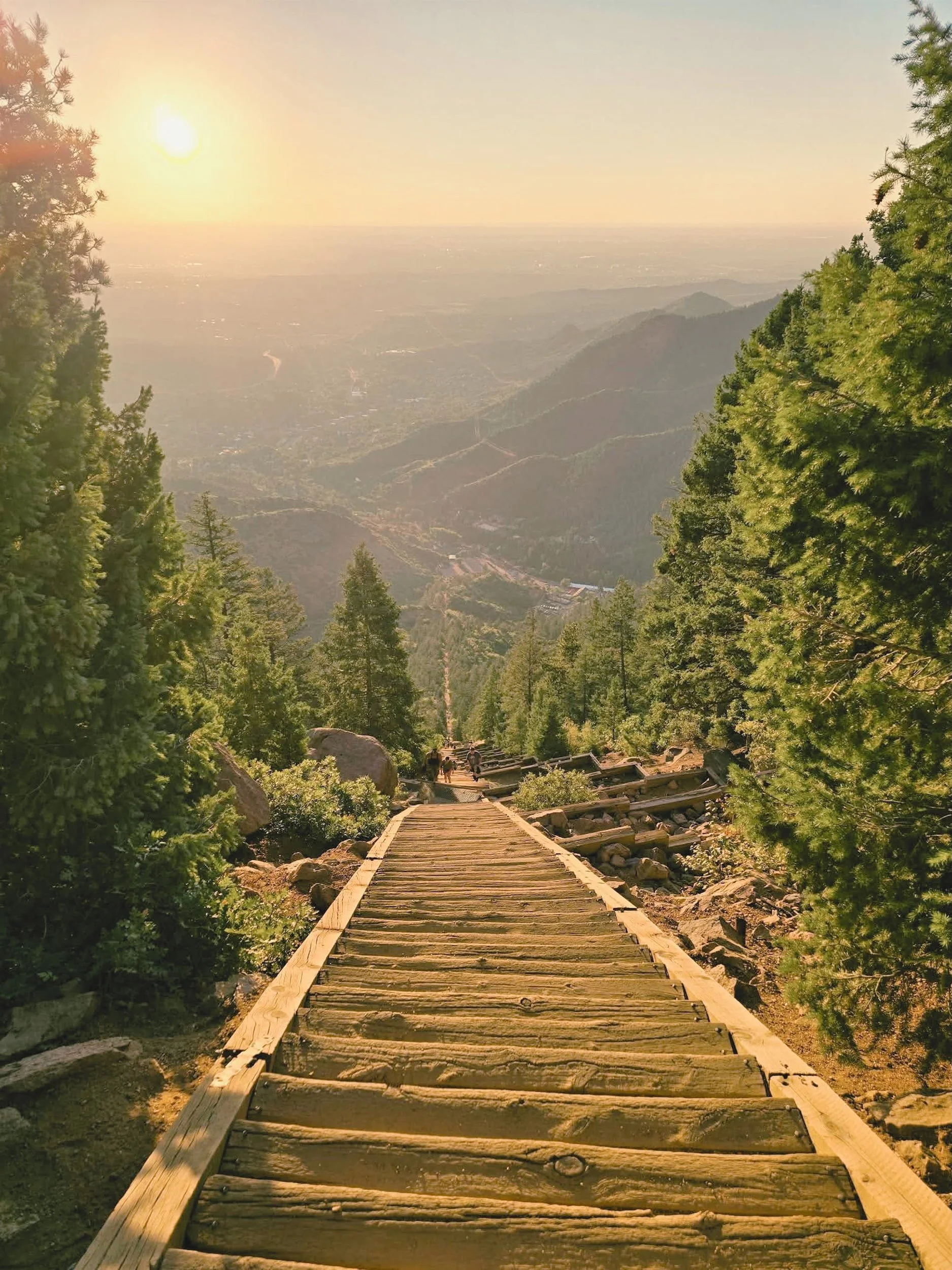 A wooden trail descending a mountain surrounded by green trees, with a view of distant valleys and the setting sun.