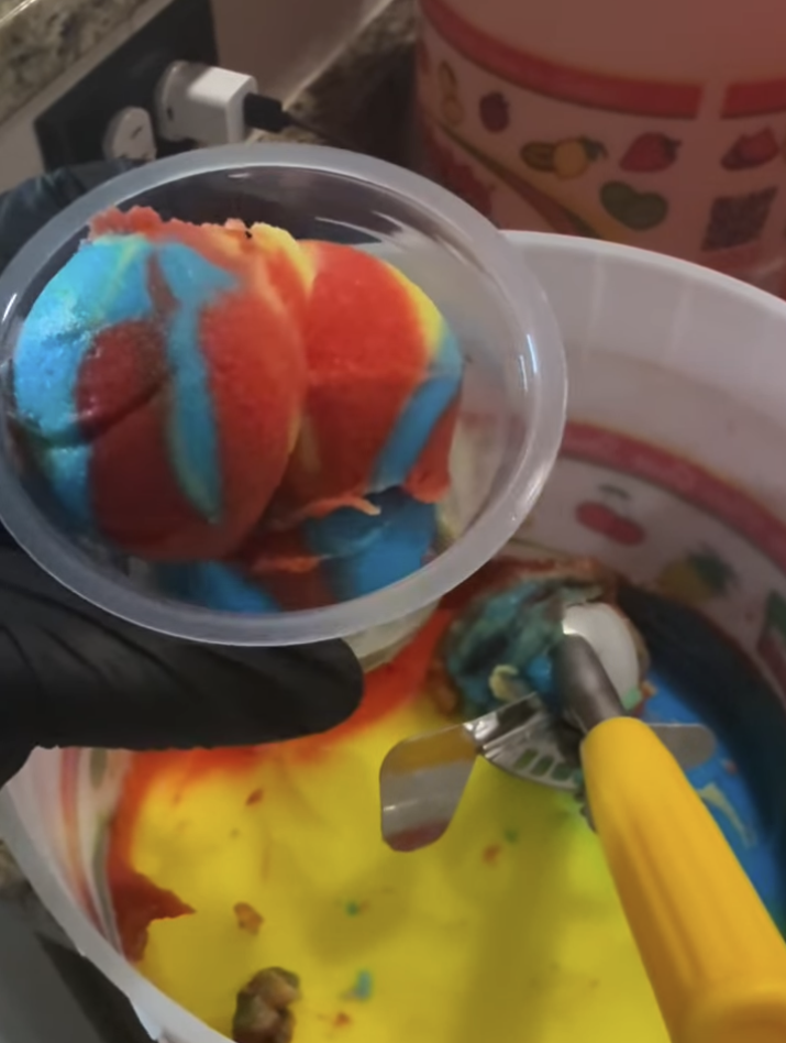 A person wearing a black glove holding a small transparent container with brightly colored, rainbow swirled ice cream or frozen dessert, next to a larger bowl of yellow and red mixture in a candy or ice cream making machine.